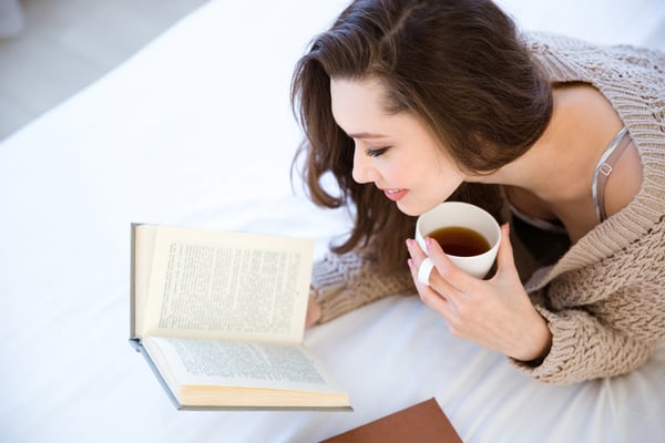 Cute lovely young woman reading book and drinking coffee on bed-1